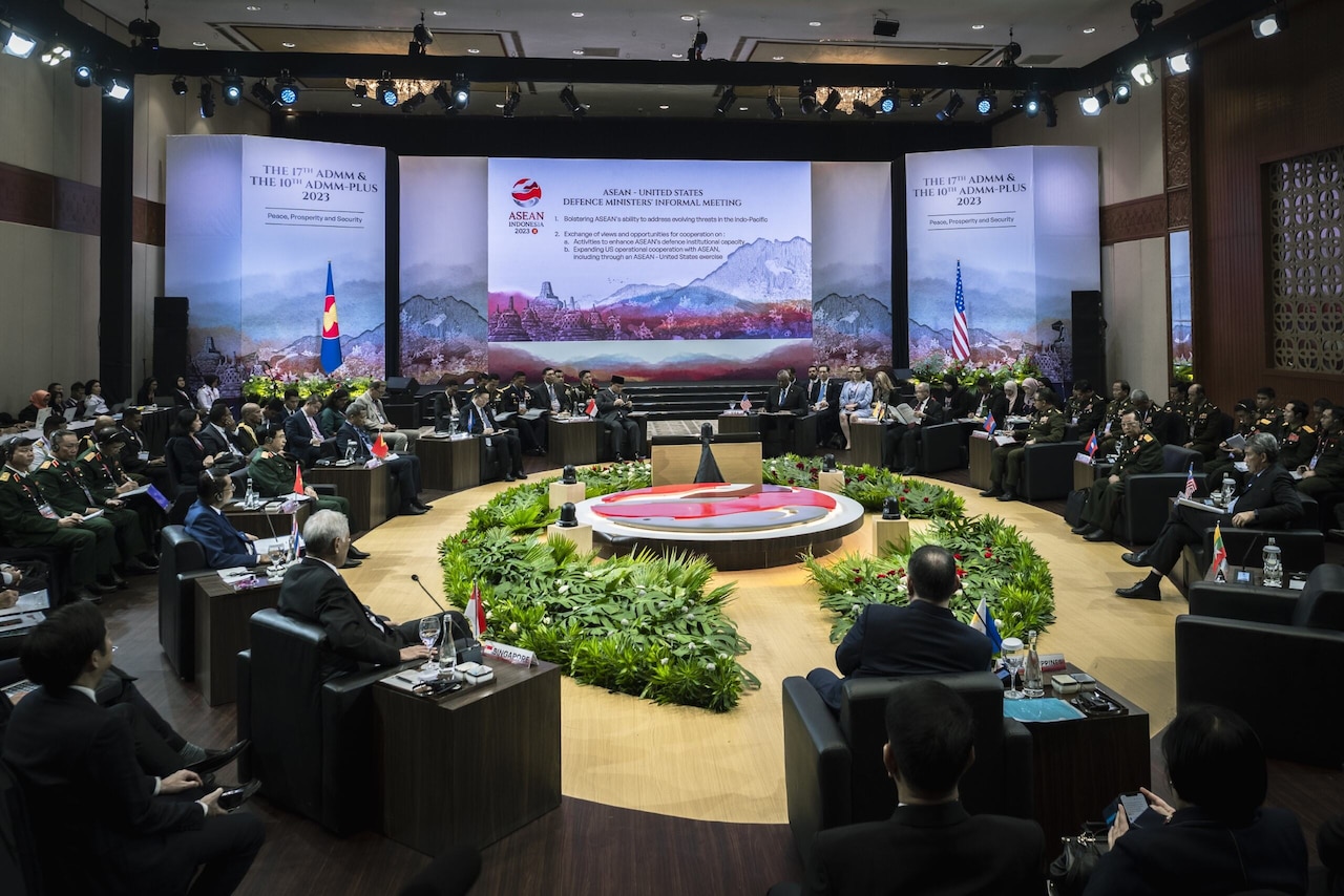 Civilians and members of foreign militaries are seated in a large meeting room.