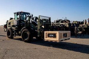 A large, wheeled military vehicle with a lifting attachment moves a crate.