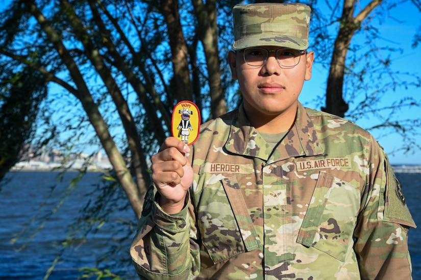 An airman in camouflage holds up a yellow oval-shaped patch while standing outdoors.