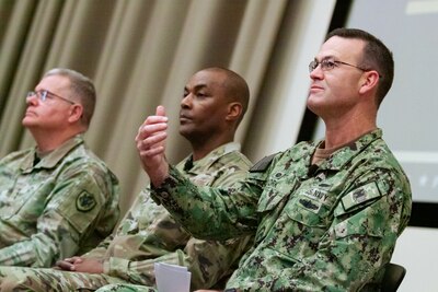 Three men in camouflage military uniforms sit on a stage. One of them is speaking and gesturing with his hand.