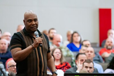 A man standing amongst an audience speaks into a microphone.