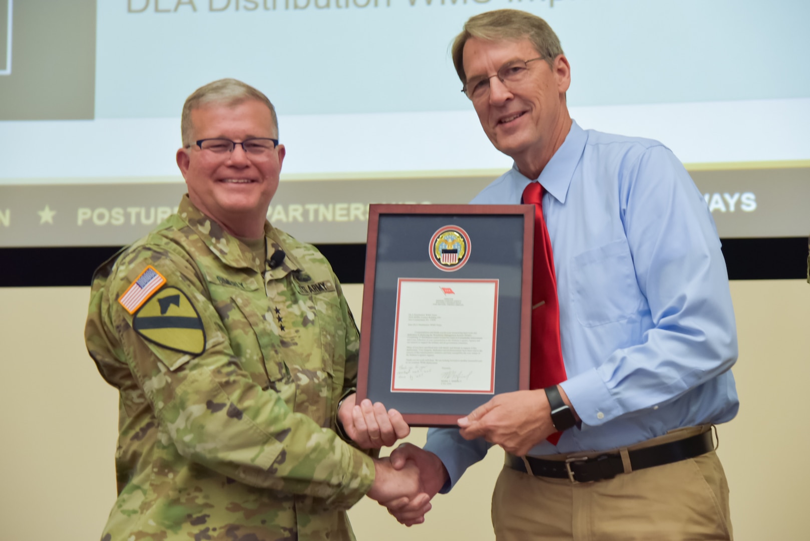A man in a camouflage military uniform shakes another man's hand. They are holding a framed certificate.