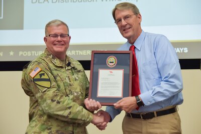 A man in a camouflage military uniform shakes another man's hand. They are holding a framed certificate.