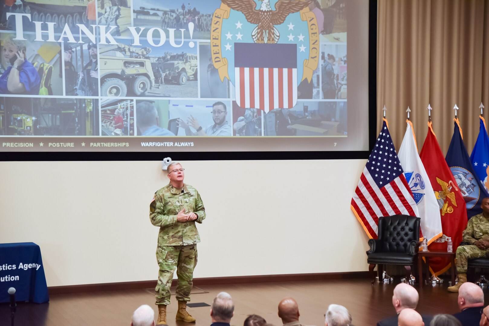 A man in a camouflage military uniform stands on stage speaking to a group of people with a screen behind him that says "Thank You!" on it.