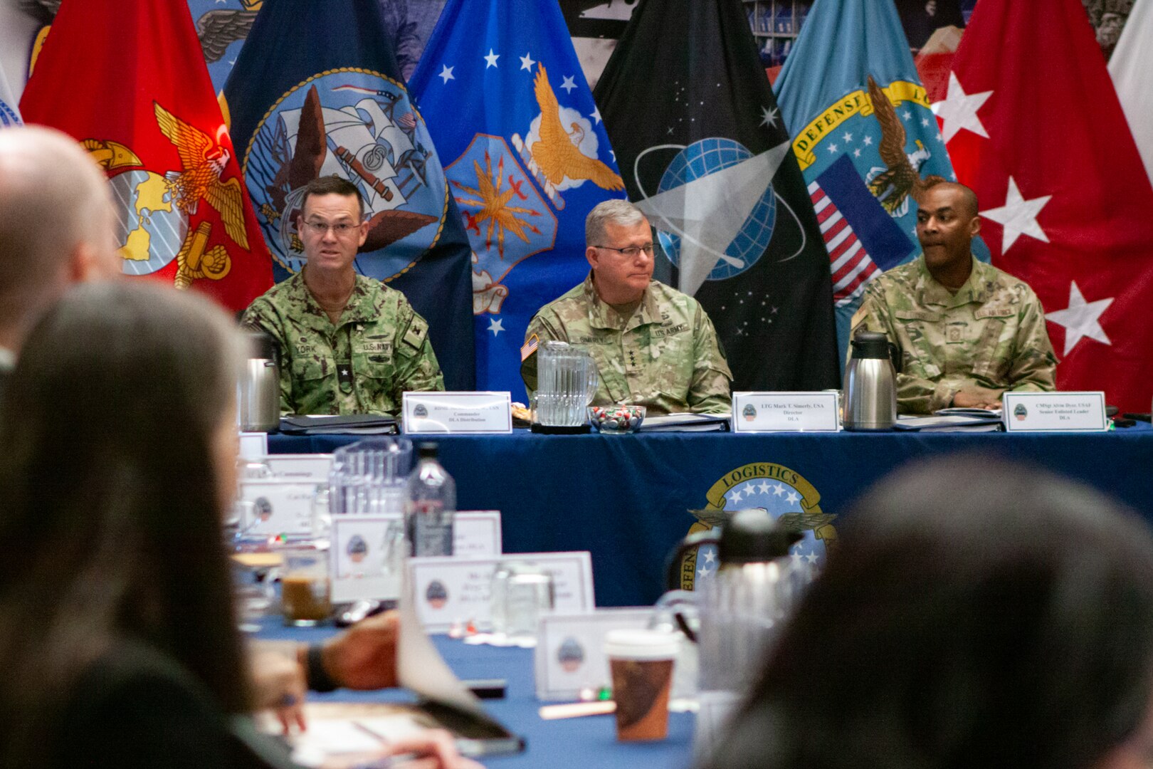 Three men in camouflage military uniforms sit at a table with flags in the background.
