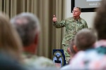 A man in a camouflage military uniform stands on stage speaking to a group of people.