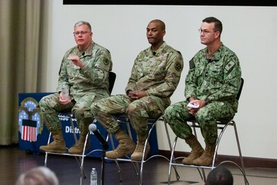 Three men in camouflage military uniforms sit on stools on a stage, one is speaking and gesturing with his hand.