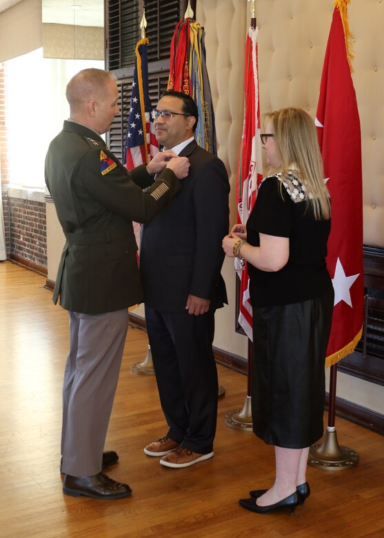 A man in an Army uniform puts a pin on the lapel of another man's suit while a woman observes.