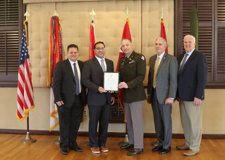 Five men pose for a photo in front of six flags. Two of the men in the center hold a certificate, one of whom is wearing the uniform of senior U.S. Army officer.