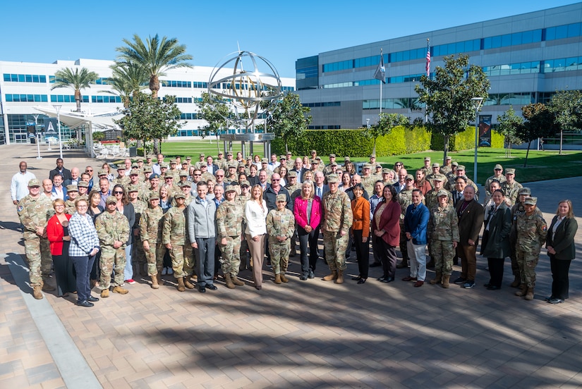 Dozens of service members and civilians stand for a group photo outside a white building.