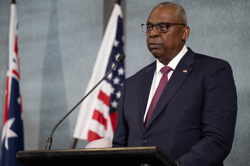 A person speaks from a podium with flags in the background.