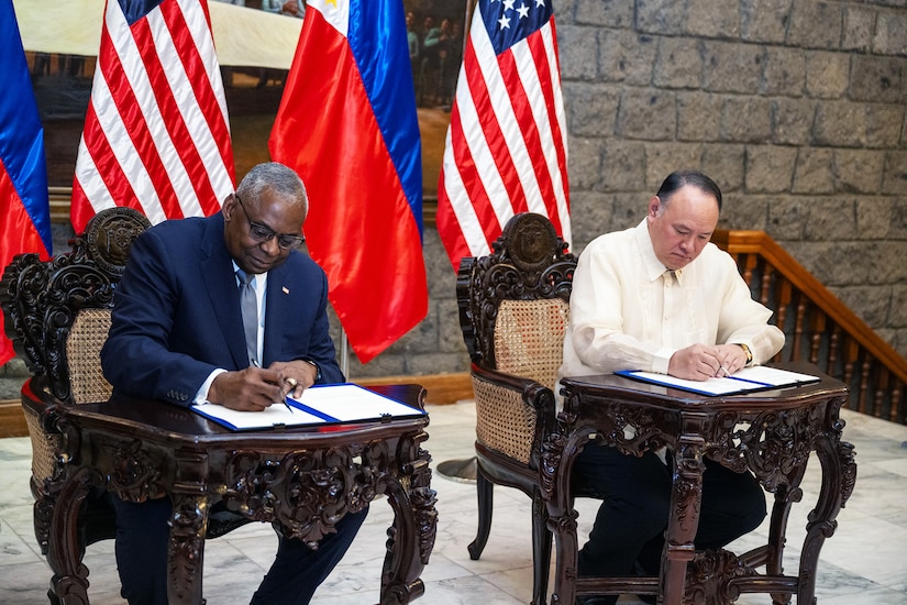 Two civilians sit at small tables and sign documents.