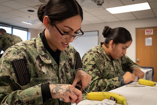 Hospitalman Elissa Segovia-Hernandez, left, and Hospitalman Cyerra Yazzie, right, practice suturing techniques on bananas during a class held aboard Naval Health Clinic Cherry Point on Friday, November 15.  Bananas served as training aids for the class due to their thick and easy-to-pierce skin and the ability to distinguish the skin from the fruit.