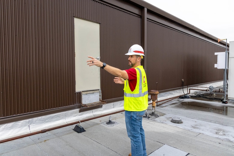 A man in blue pants, a red shirt, yellow safety vest and white hardhat points to the left in front of a brown wall and stands on a grey floor.