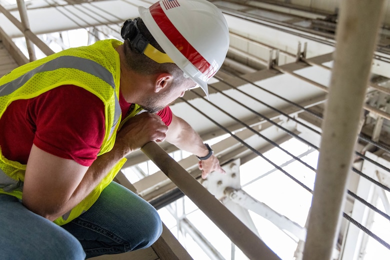 A man in a red shirt, yellow safety vest, and white hardhat points at grey beams.
