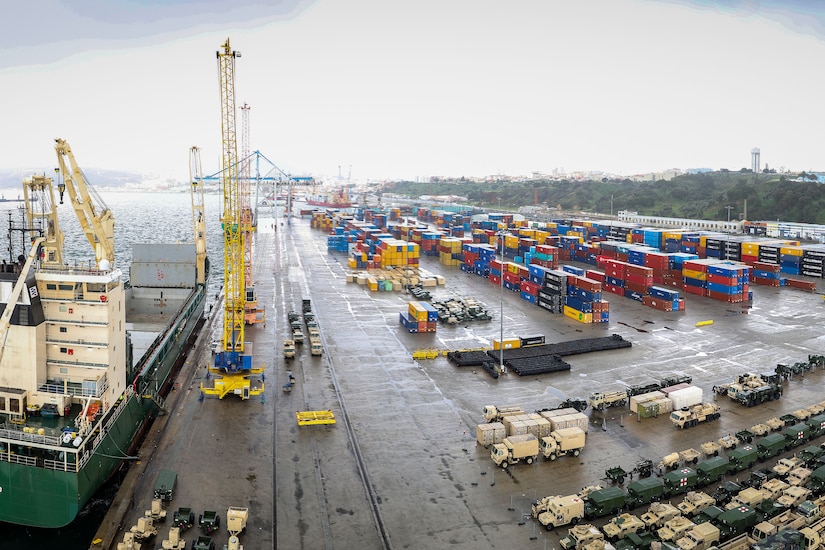 Vehicles and containers are staged at a port, where a yellow crane sits near a vessel.