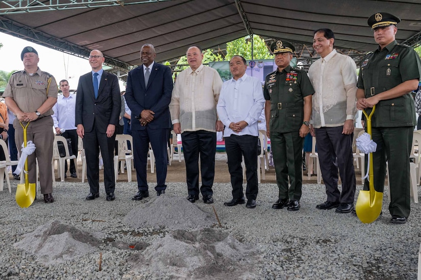 Two men wearing U.S. and foreign military uniforms hold shovels while standing next to men wearing civilian clothes.