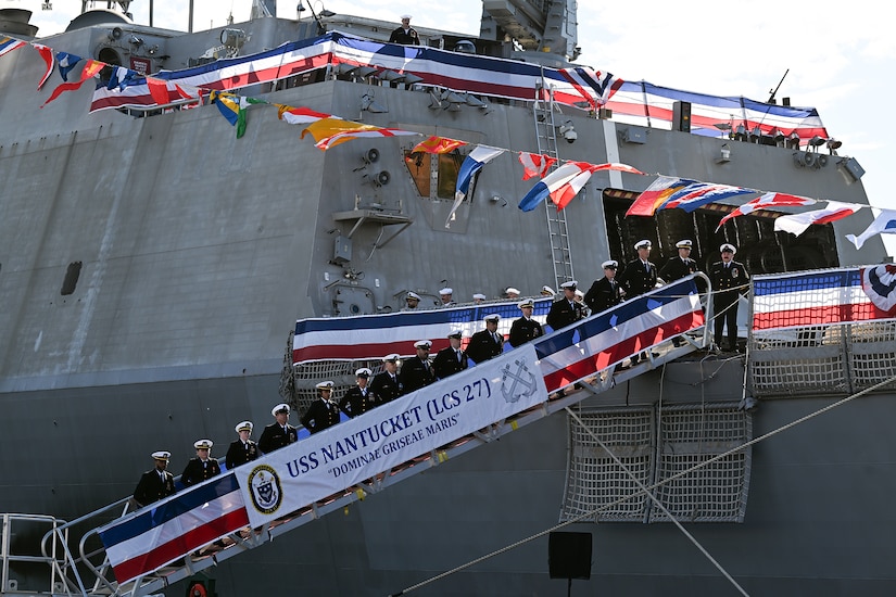 Sailors stand in row on a red, white and blue banner-festooned ramp to a ship.