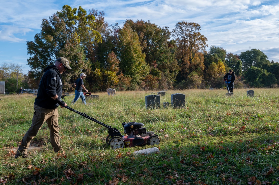 Volunteer pictured mowing the grass with other volunteers in the background and graves.