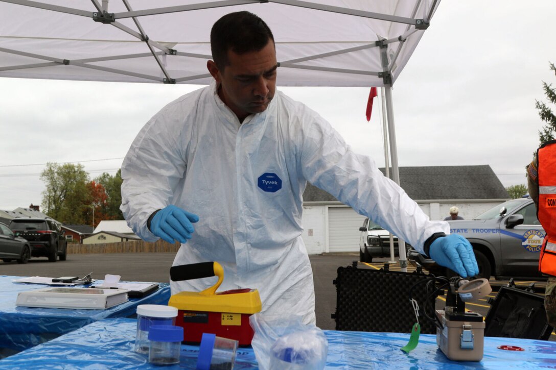 A man wearing coveralls and rubber gloves uses forensics equipment arrayed on a table.