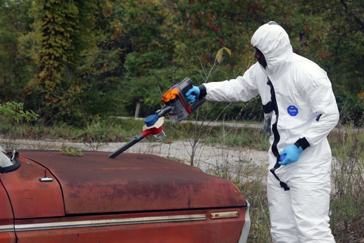 A man in a hazardous material suit vacuums particles off a vehicle.