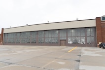 A red brick building with a wall full of windows sits on a brown concrete pad with a grey sky in the background.
