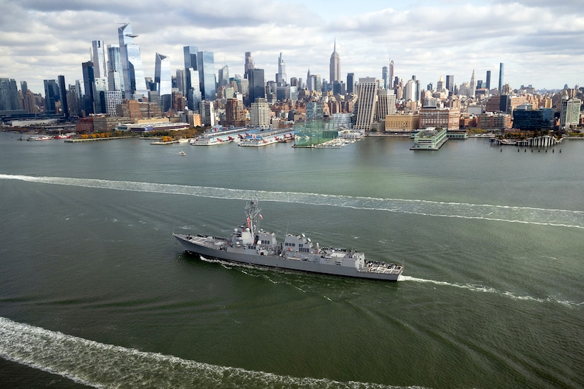 A military ship sails next to the New York City coastline on a river.