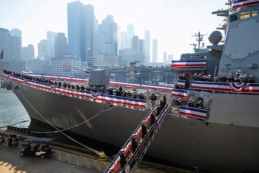 A large military ship adorned with blue, white and read ribbons sits at port with sailors on deck.