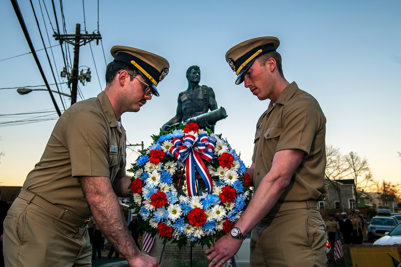 A pair of sailors hold a wreath in front of a statue.