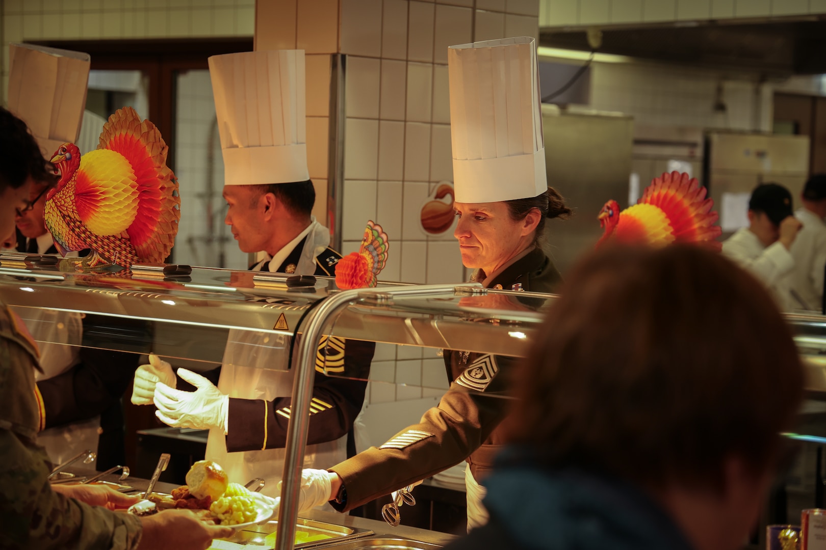 U.S. Army Command Sgt. Maj. Amanda Reuter, senior enlisted advisor, 21st Special Troops Battalion, 21st Theater Sustainment Command, hands a plate of food to a Soldier while serving Thanksgiving meals to Soldiers and families at the Clocktower Cafe on Kleber Kaserne, Kaiserslautern, Germany, on Nov. 22, 2023.