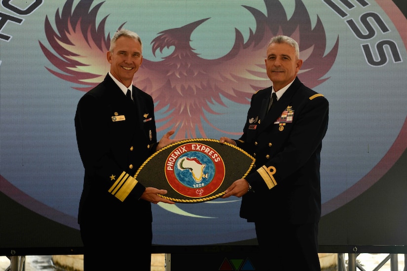 Two high-ranking navy officials hold a seal in front of a logo of a phoenix.