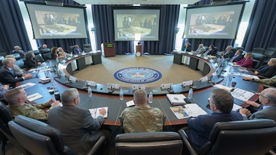 Over a dozen men and women sit at a circular table facing three wall monitors.