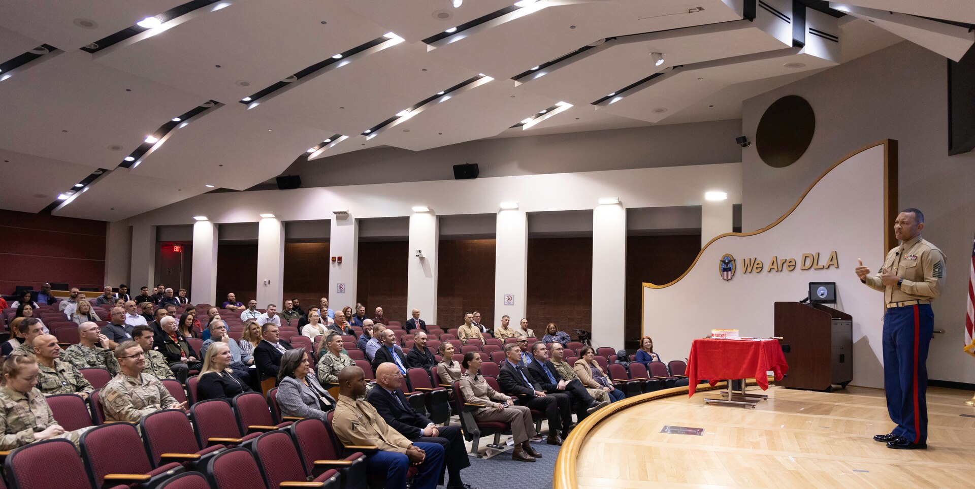 A dark skinned man stands on stage and speaks in front of a audience in an auditorium. He is wearing a Marine Corps tan uniform with a tan shirt and blue pants.
