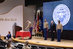 Five people stand on a stage. One is in a military uniform and is cutting a cake. The other three men are standing at attention on the stage. Two men flank the middle man who is not in uniform, rather in a business suit.