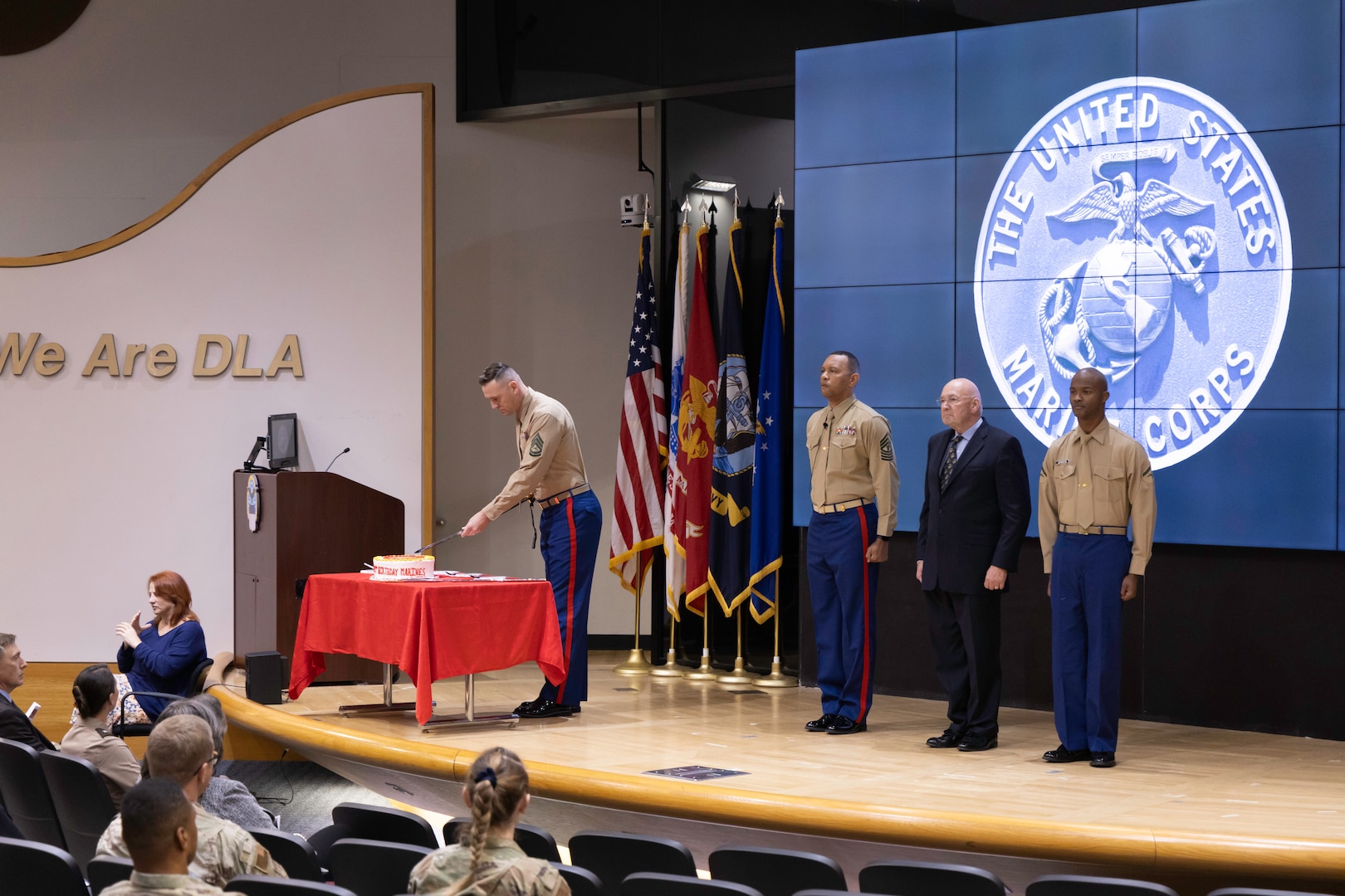Five people stand on a stage. One is in a military uniform and is cutting a cake. The other three men are standing at attention on the stage. Two men flank the middle man who is not in uniform, rather in a business suit.