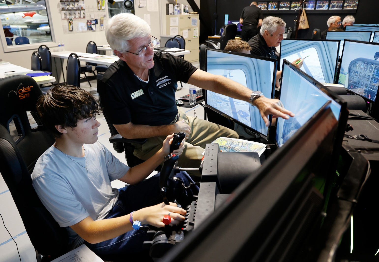 A student sits at a flight simulator station with a large screen showing an in-flight view, hands on the controls, while a volunteer instructor beside them offers guidance. Other simulator stations with students and instructors are visible in the background, showing a full classroom engaged in pilot training.