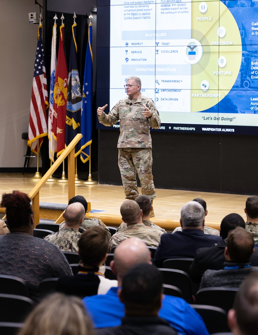 A light skinned man with graying hair in uniform speaks in front of an audience in a auditorium.