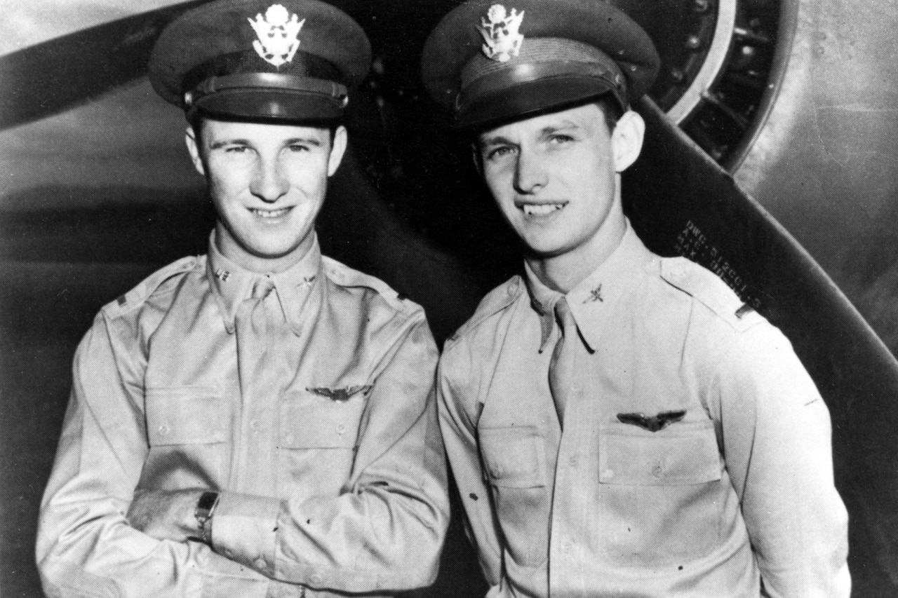Two pilots stand in front of a parked aircraft and smile for a photo.