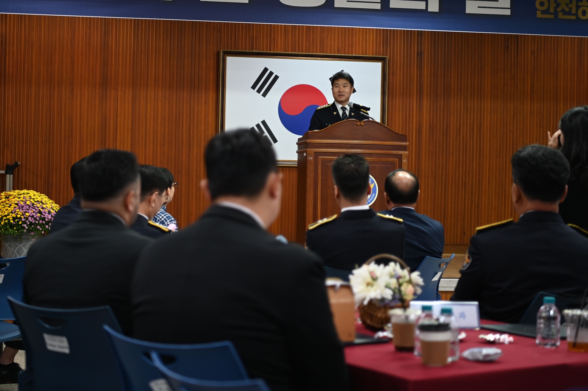 A man stands on stage in front of the Republic of Korea's flag behind a podium in front of an audience. They are all wearing formal police attire or suits.