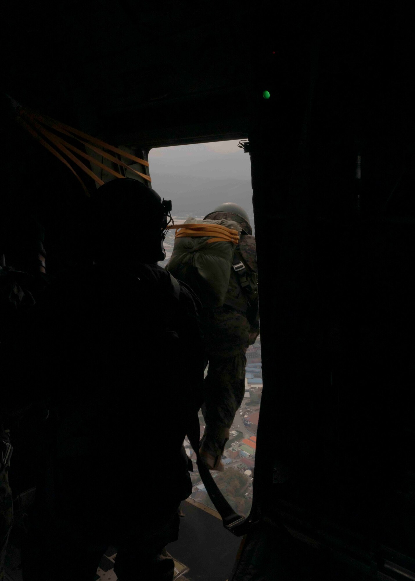 A paratrooper jumps out of a plane's side door, silhouetted by a black door.