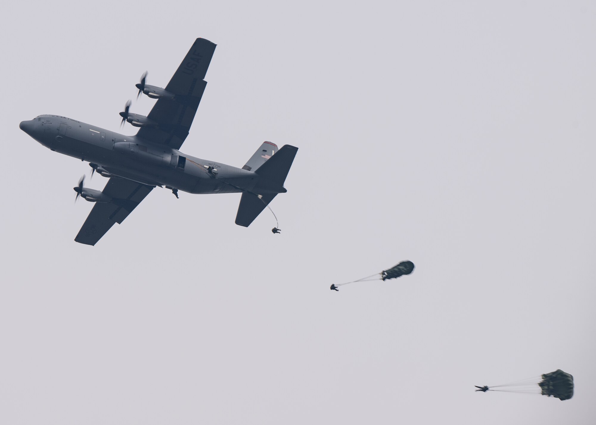 A gray C-130J cargo airplane with four propellers fly to the left in a gray sky.
