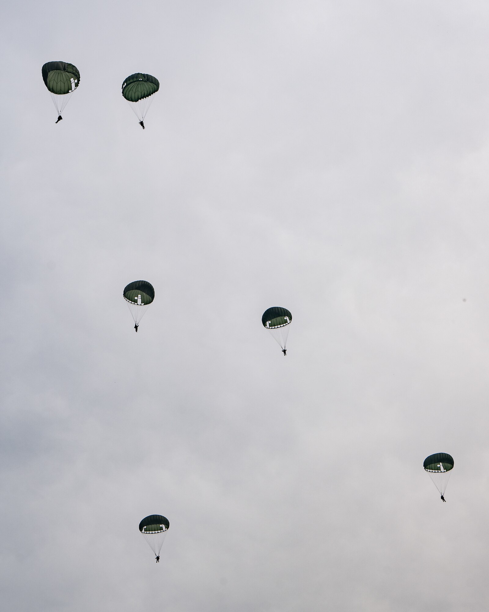 Six men hang from deployed circular parachutes against a cloudy gray sky.