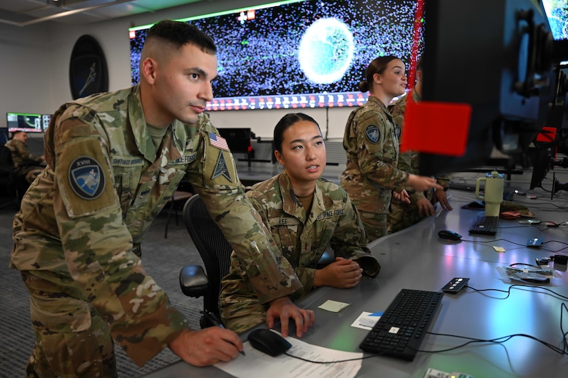 Two guardians in front of a long desk look closely at a monitor as other guardians work in the room around them.