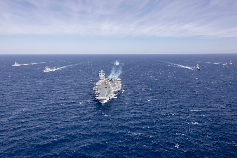 A group of military ships steam in formation in the ocean under a partly cloudy sky.