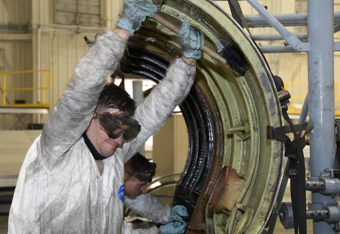 Photo shows two men with large brushes scrubbing an aircraft part.