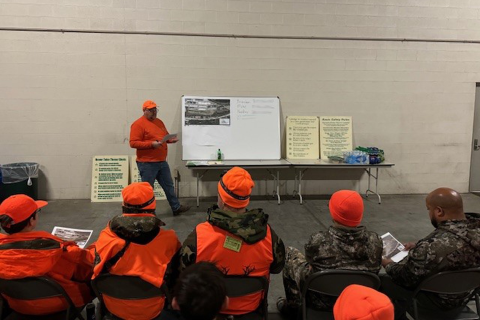 A man in an orange shirt and hat stands talking in front of a seated group wearing orange hunting vests and camouflage.