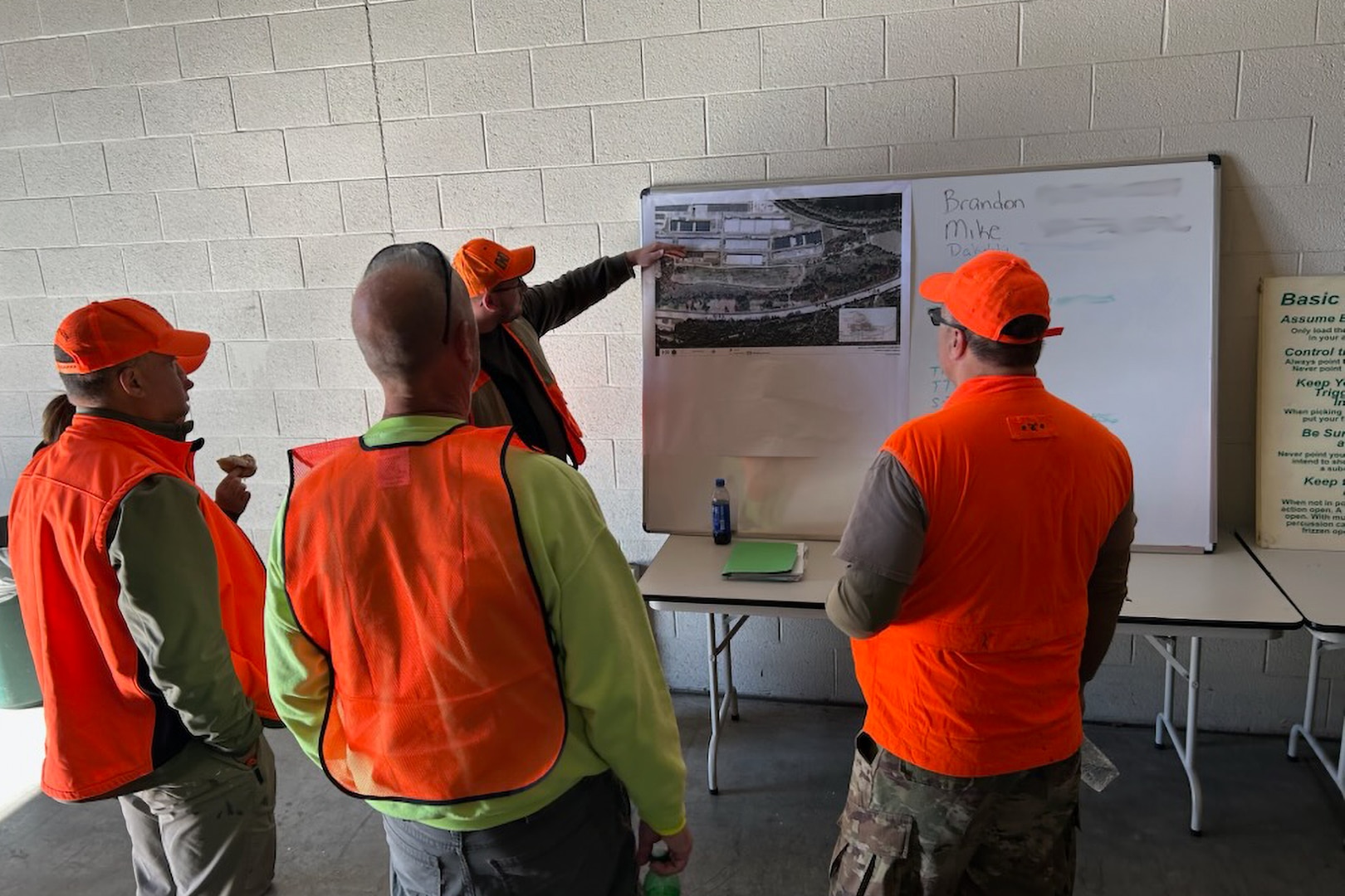 Four men in orange hunting vests stand around a whiteboard with a map hanging on it. One man is pointing at the map.