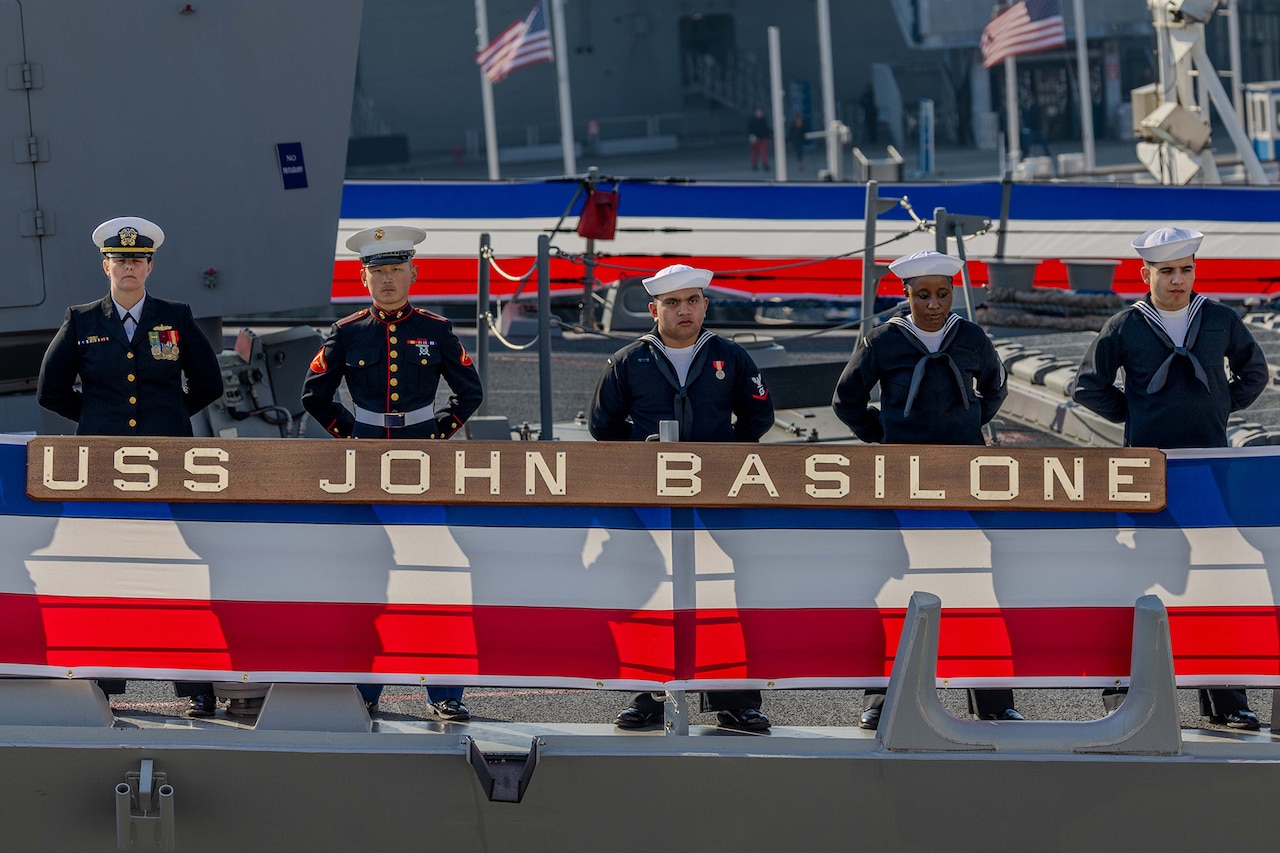 Sailors stand at attention by the rails on the deck of a U.S. naval ship.