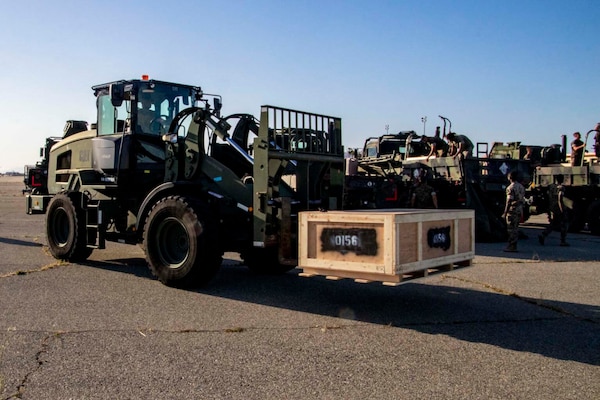 A large, wheeled military vehicle with a lifting attachment moves a crate.