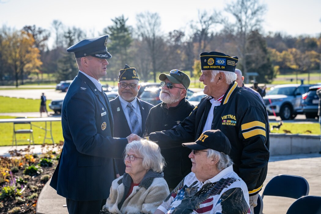 Col. Richards shakes hands with an older veteran wearing a black and gold Vietnam veteran jacket and flight cap adorned with pins